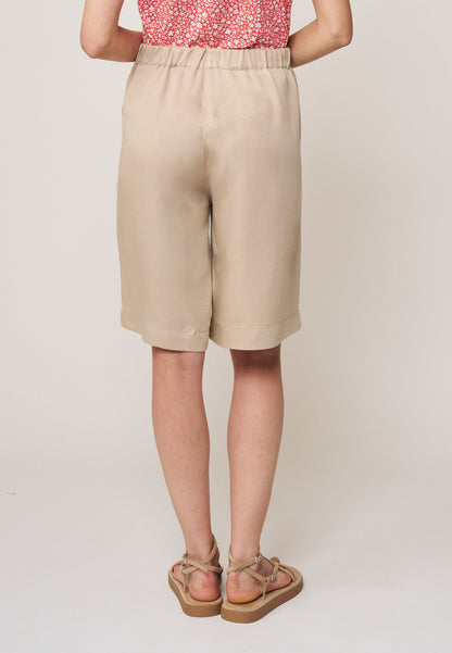 Frau von hinten in hellen, locker geschnittenen Shorts, Modell Azita, mit elastischem Bund und Sandalen vor hellem Hintergrund